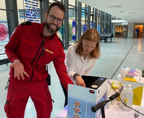 Person ikledd rød uniform med "Ambulanse" på har hånden i en håndhygienemaskin og får hjelp av ansatt fra Kiilto og Antibac. Innendørs (foajee).