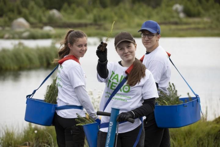 Tre unge personer fra 4H planter trær som del av Kiilto sitt miløinitiativ. De står ved et vann, og har på seg hvite t-skjorter, med stiklingene i blå baljer.