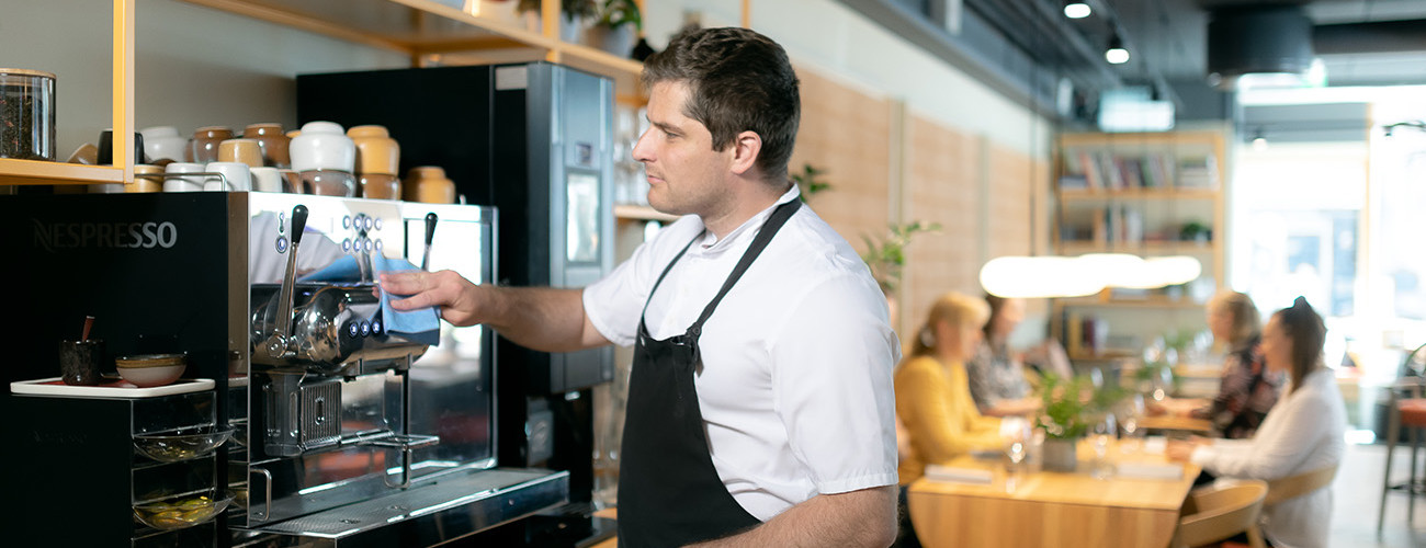 Barista foran en espressomaskin i kafé, glade kunder ved bord i bakgrunnen.